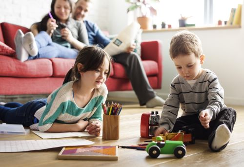 Boy and girl playing on hardwood floor with parents watching from couch in clean home.